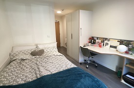 A room with a double bed in one corner and a desk and wardrobe against the opposite wall. The desk has an office chair next to it and shelves beneath it.
