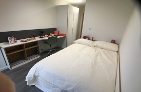 A room with a double bed in one corner and a desk and wardrobe against the opposite wall. The desk has an office chair next to it and shelves beneath it.