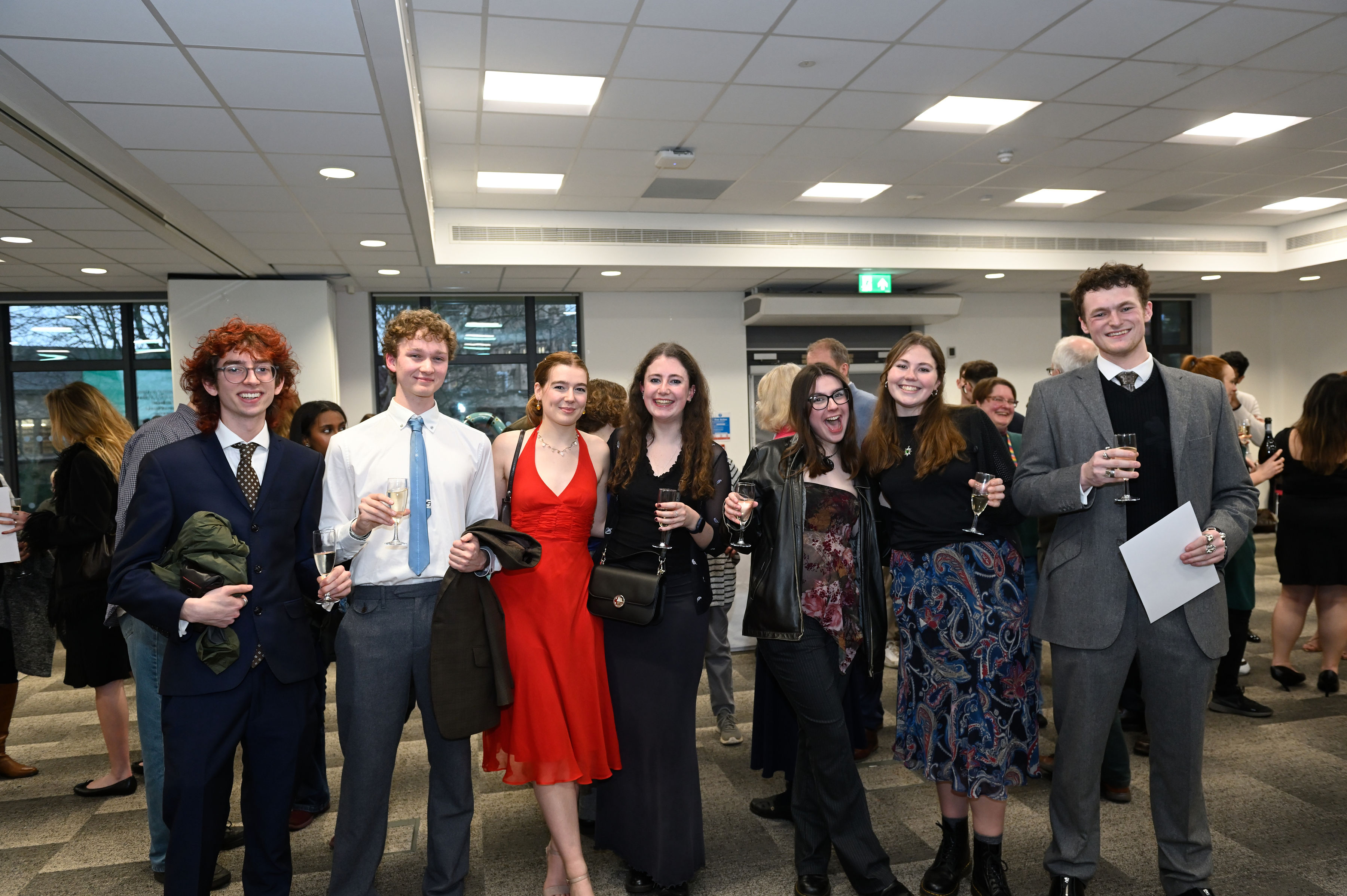 Group of 7 law student prize winners, stood in a row, smiling and looking at the camera, wearing smart clothes and holding champagne flutes.